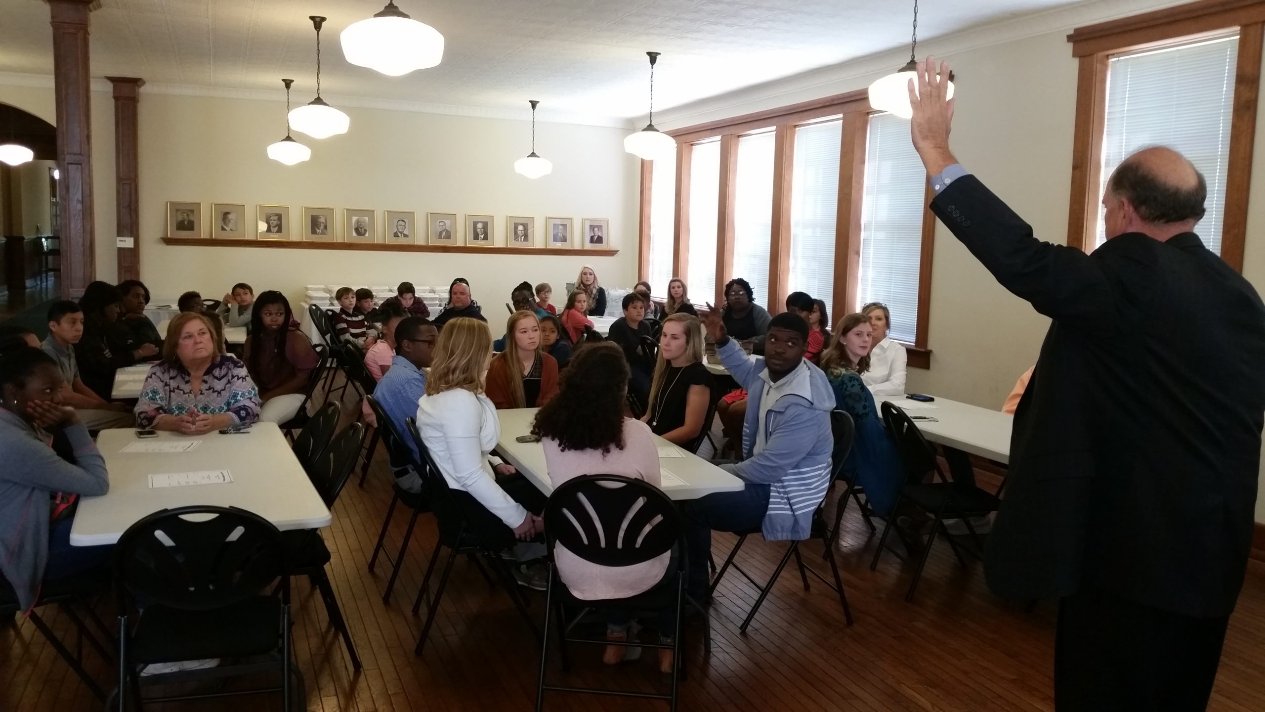 Crowd of students at tables with Dr. Miller speaking and his hand raised